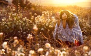 Junges Mädchen mit langen blonden Haaren, sitzt in einem wolligen grauen Poncho auf einem Feld, mit weißen Blumen während eines Sonnenuntergangs.