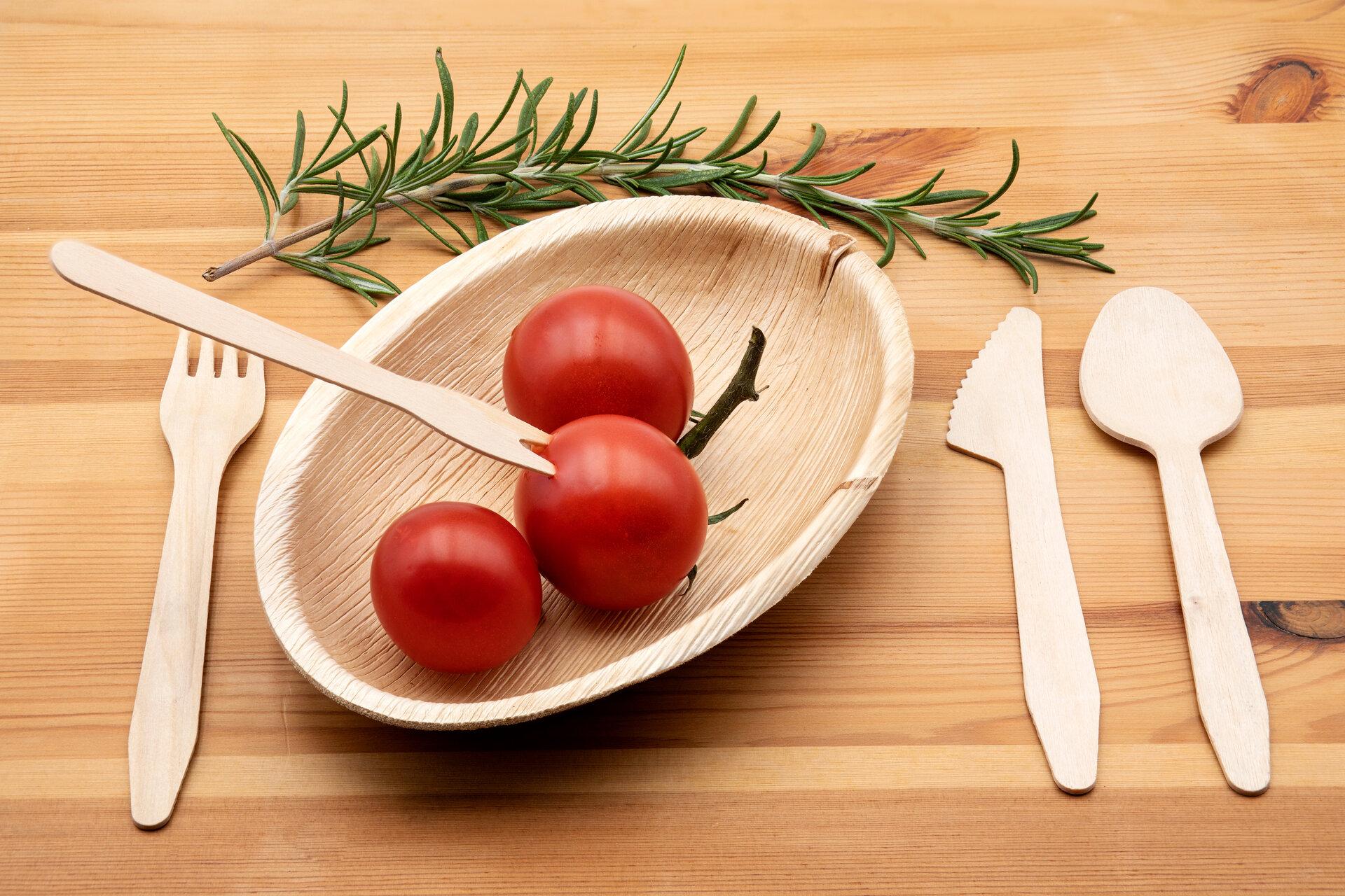Hölzernes Einweg-Besteck auf einem Holzbrett mit Tomaten und Rosmarin.